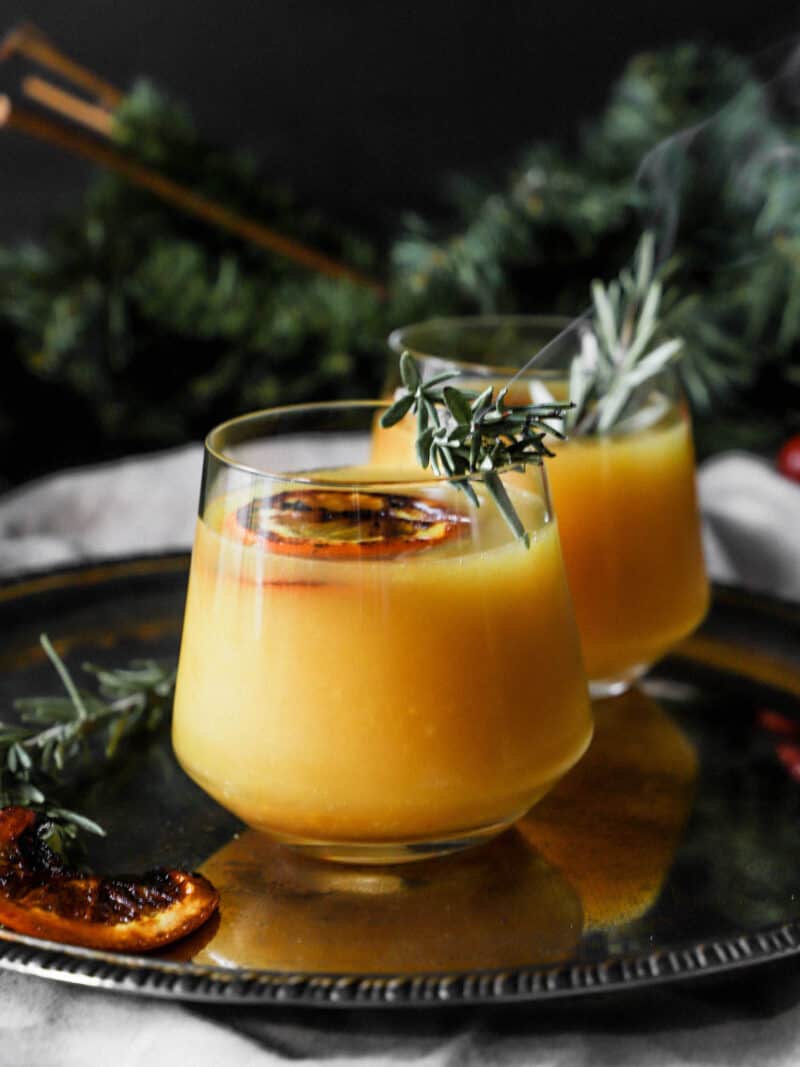 two glasses of a bruleed orange old fashioned cocktail on a serving platter in front of holiday decor