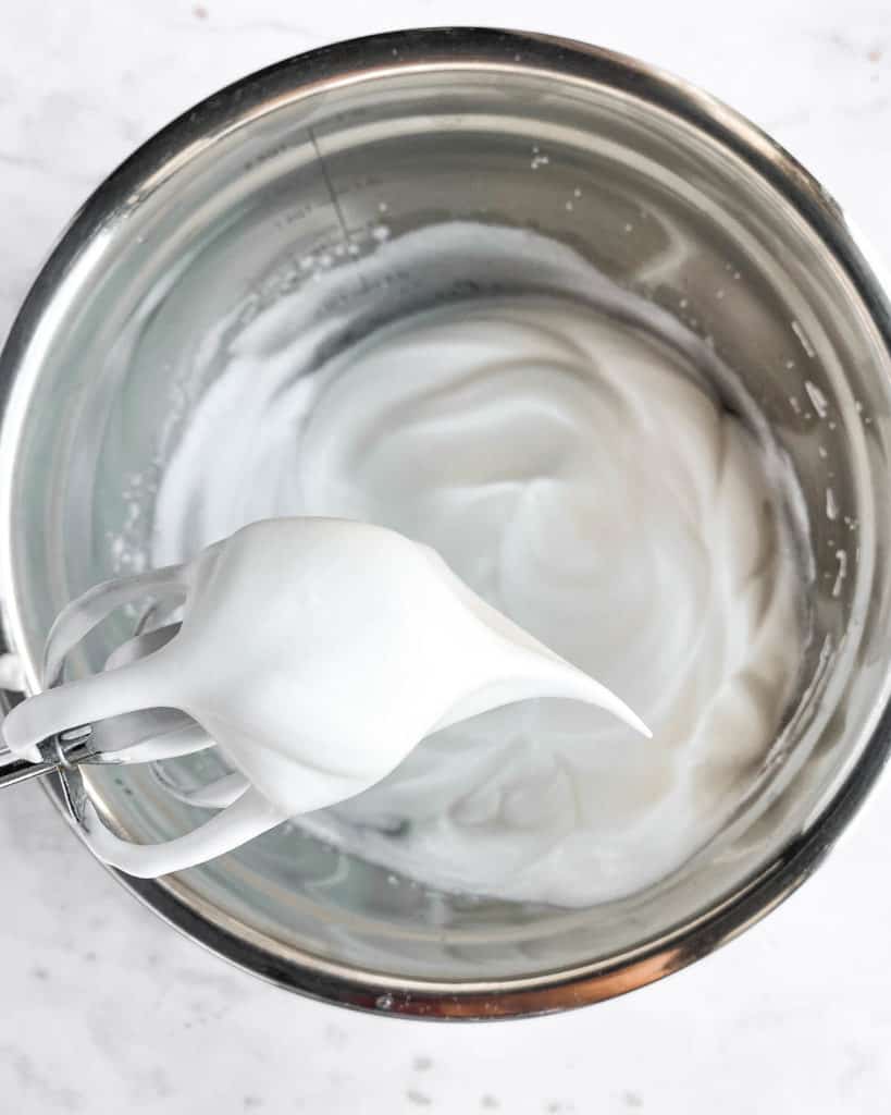 whipped meringue in a bowl with a soft peak on the mixer whisk